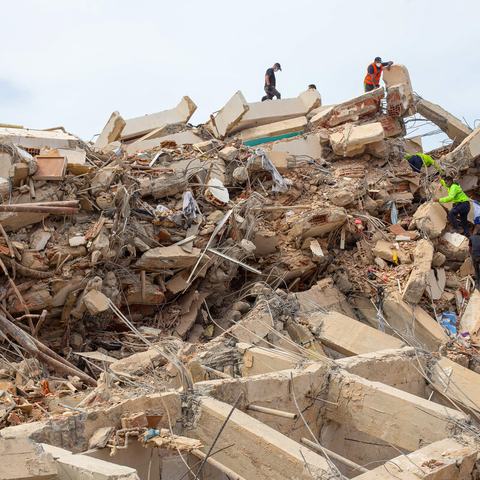 Rescue workers climb over the remains of a collapsed concrete structure, searching through piles of rubble, broken beams and exposed steel reinforcement.