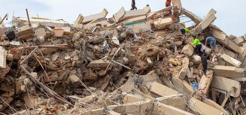 Rescue workers climb over the remains of a collapsed concrete structure, searching through piles of rubble, broken beams and exposed steel reinforcement.