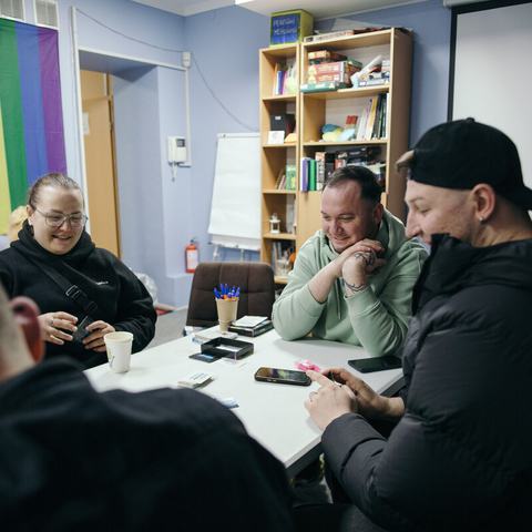 Four people sat around a table in an office decordated with rainbow coloured blinds at the window. The people at the table at smiling and having a conversation.