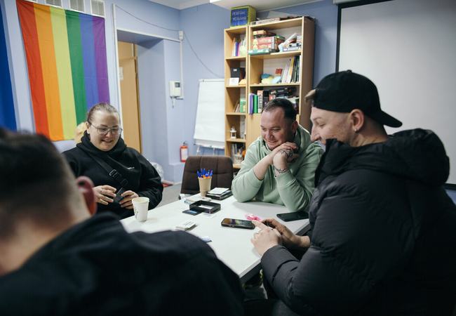 Four people sat around a table in an office decordated with rainbow coloured blinds at the window. The people at the table at smiling and having a conversation.