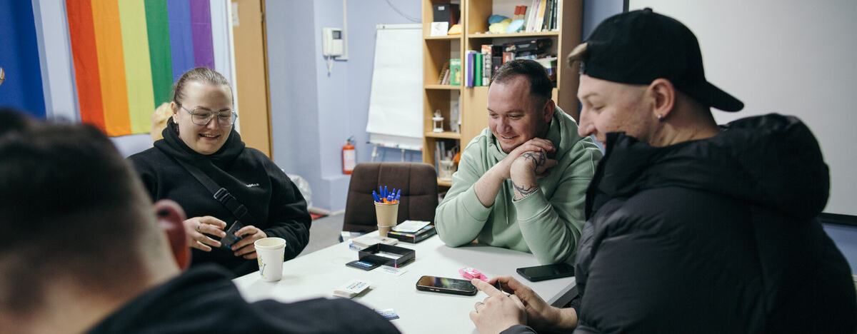 Four people sat around a table in an office decordated with rainbow coloured blinds at the window. The people at the table at smiling and having a conversation.