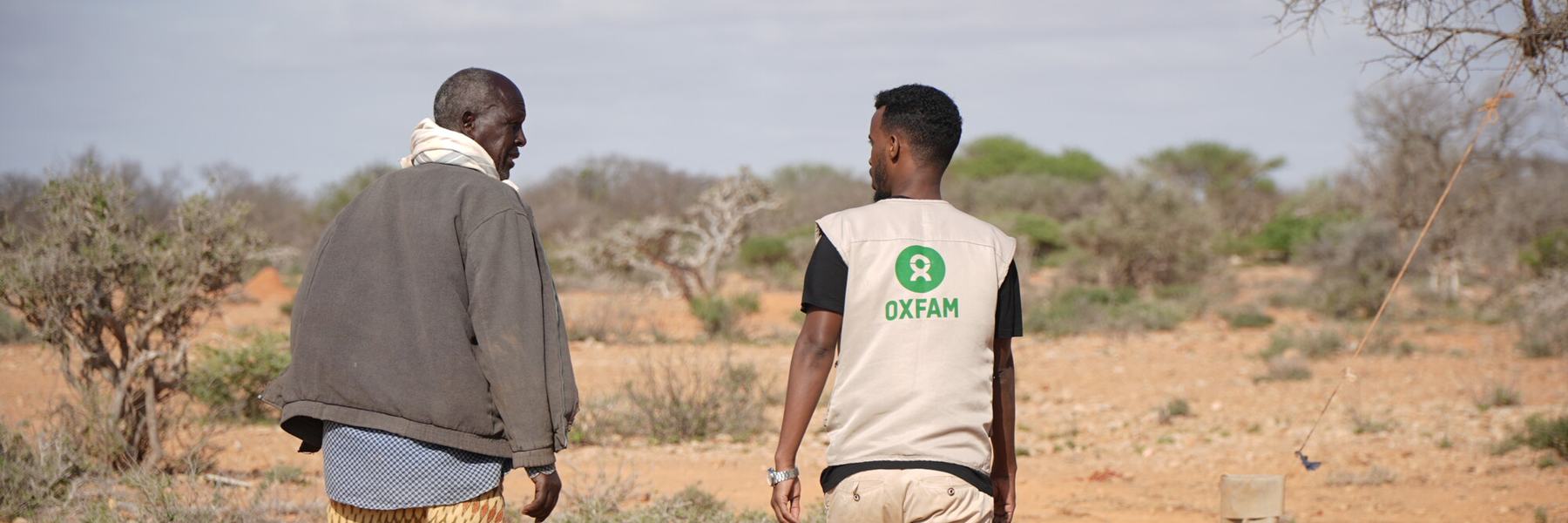 Two men, one wearing an Oxfam-branded vest, walk through a dry, drought-ridden landscape.