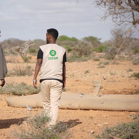Two men, one wearing an Oxfam-branded vest, walk through a dry, drought-ridden landscape.