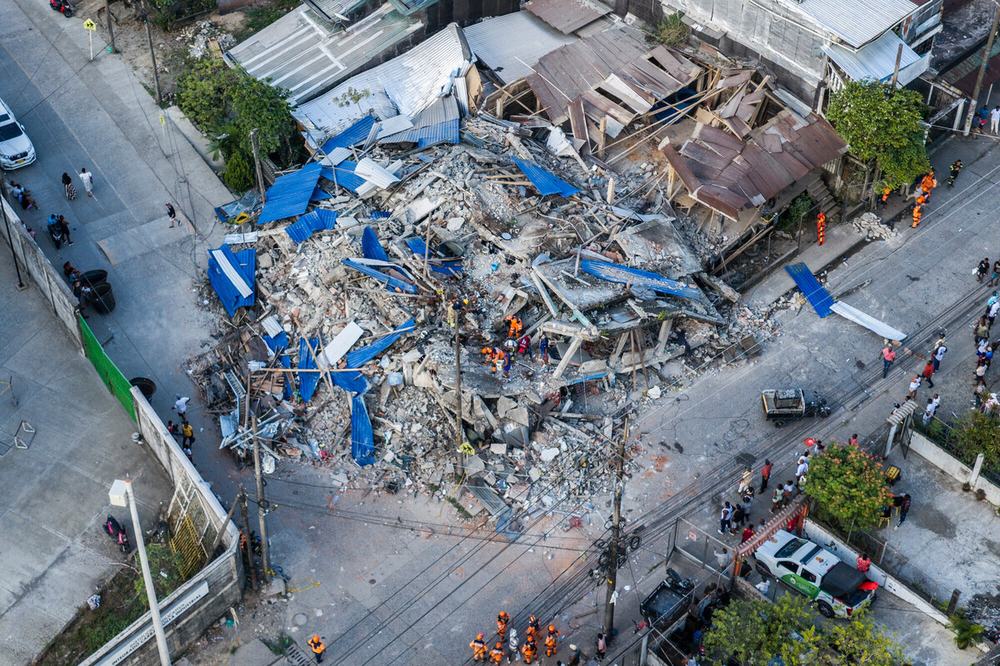 Aerial view of an earthquake-affected area showing collapsed buildings, debris, and emergency teams conducting search and response operations. The image highlights the scale of the damage across the territory.