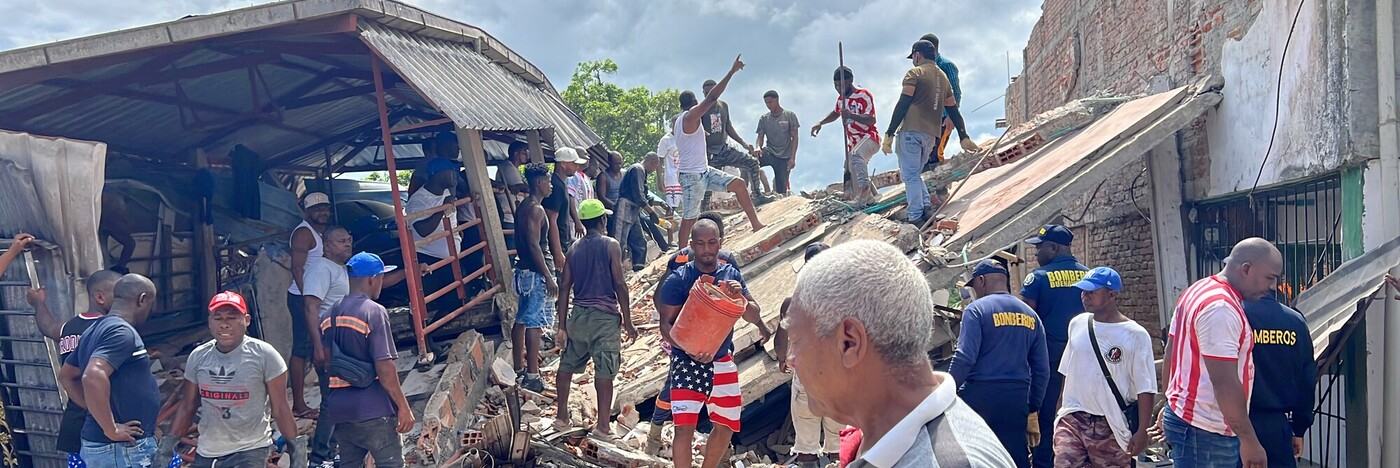 Members of the community and emergency response teams helping to clear debris and rubble from a collapsed building. People are carrying buckets and climbing over the structure to assist in the clear up.
