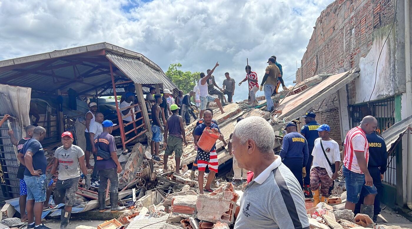 Members of the community and emergency response teams helping to clear debris and rubble from a collapsed building. People are carrying buckets and climbing over the structure to assist in the clear up.