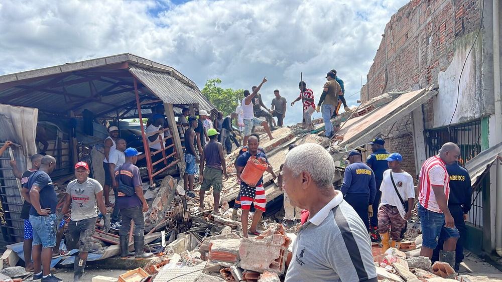 Members of the community and emergency response teams helping to clear debris and rubble from a collapsed building. People are carrying buckets and climbing over the structure to assist in the clear up.