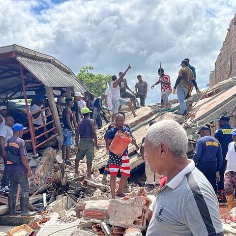 Members of the community and emergency response teams helping to clear debris and rubble from a collapsed building. People are carrying buckets and climbing over the structure to assist in the clear up.