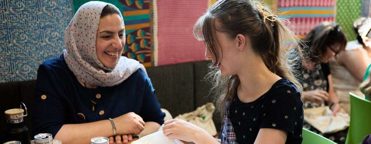 Two women are sat at a table. One of the women is doing some embroidery and the other woman is watching and smiling.