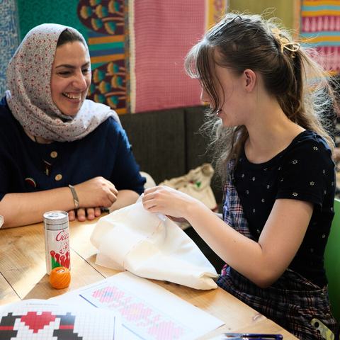 Two women are sat at a table. One of the women is doing some embroidery and the other woman is watching and smiling.