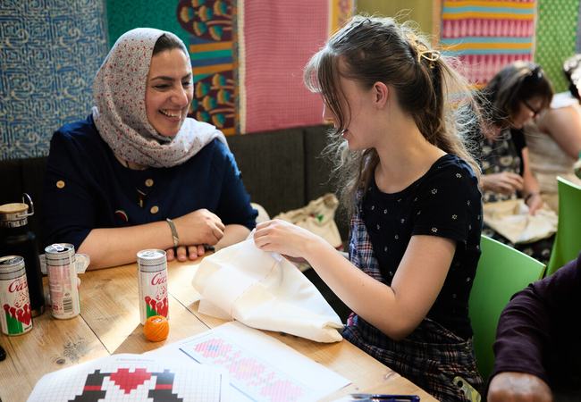Two women are sat at a table. One of the women is doing some embroidery and the other woman is watching and smiling.