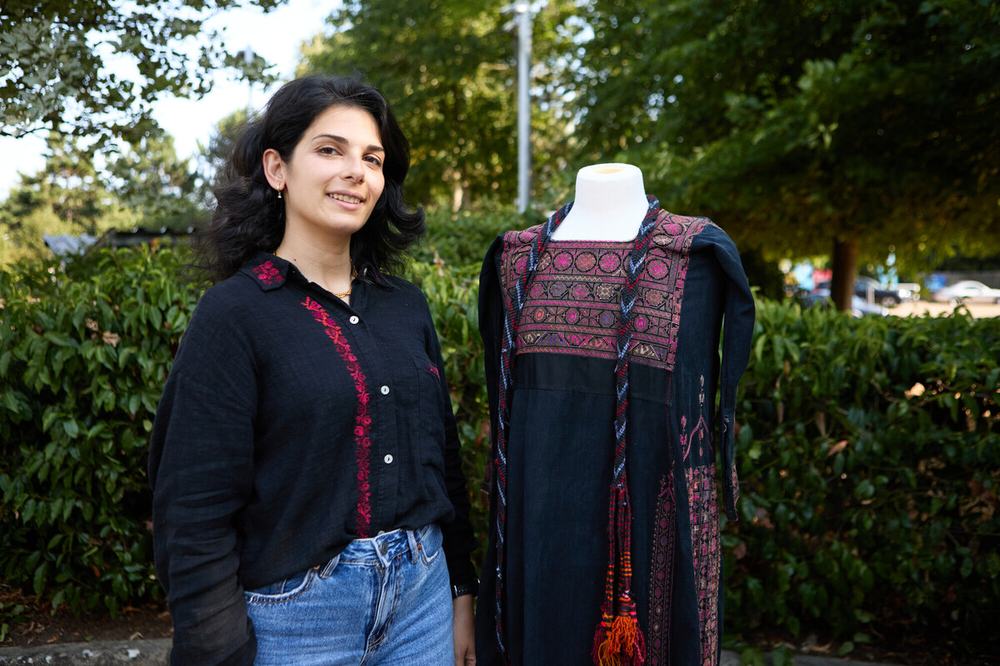 Dina Asfour of The Tatreez Institute is stood outside next to a pre-Nakba thobe in black with pink, orange and blue embroidery known as tatreez.