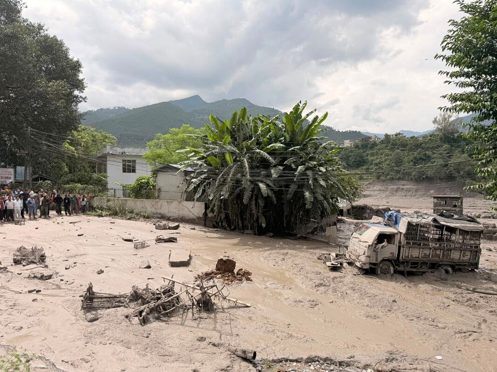 A group of people are stood together near a house looking at the flood water in the road and a damaged truck.