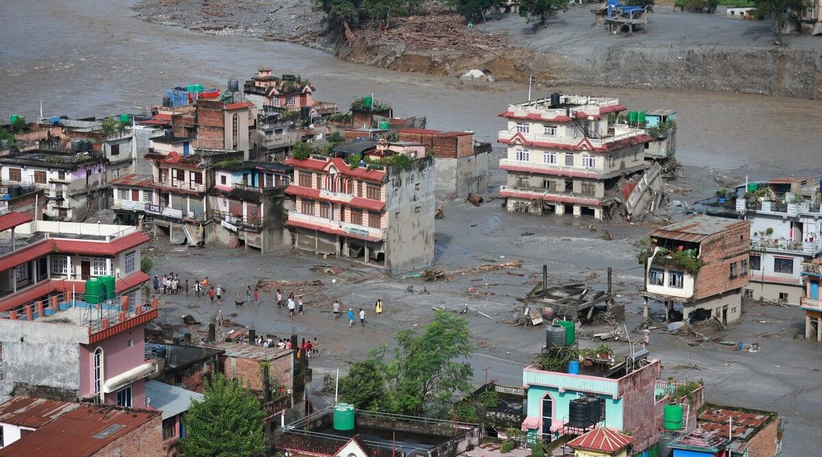 Buildings surrounded by muddy flood water. People are stood in the deep water looking at the buildings destroyed by the floods.