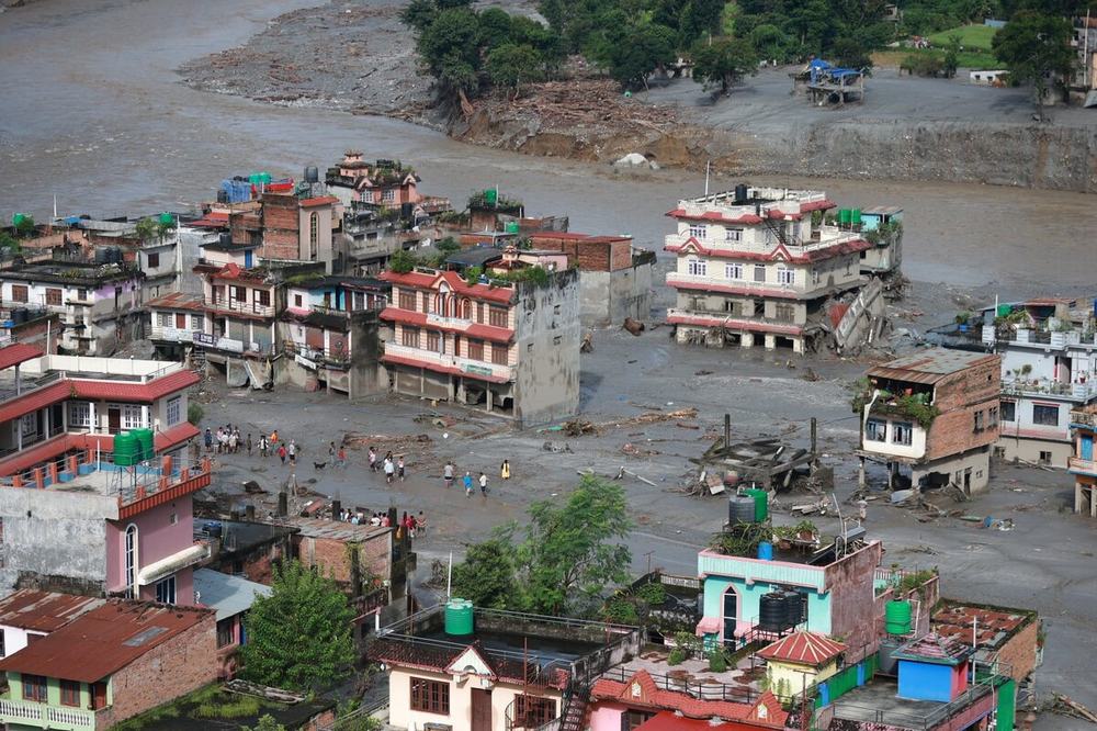 Buildings surrounded by muddy flood water. People are stood in the deep water looking at the buildings destroyed by the floods.