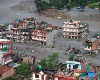 Buildings surrounded by muddy flood water. People are stood in the deep water looking at the buildings destroyed by the floods.