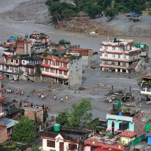 Buildings surrounded by muddy flood water. People are stood in the deep water looking at the buildings destroyed by the floods.
