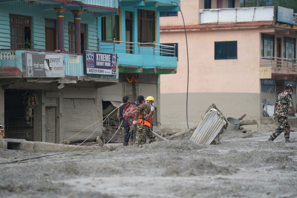 A group of people are stood in the mud near some damaged buildings in the aftermath of the landslides in Bidur Muncipality, Nuwakot District, Nepal.