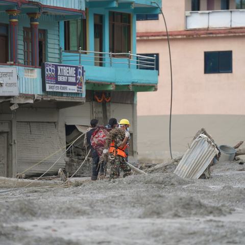 A group of people are stood in the mud near some damaged buildings in the aftermath of the landslides in Bidur Muncipality, Nuwakot District, Nepal.