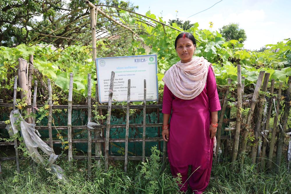 A woman stands in front of a fence. Behind the fence, is a farm tunnel in which plants are growing.