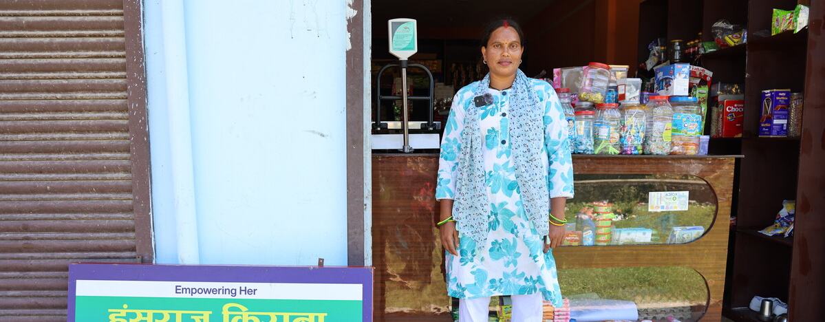 A person stands at the entrance of a local shop stocked with groceries and household goods, beside a sign advertising the business.