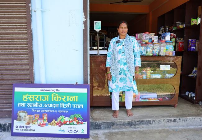 A person stands at the entrance of a local shop stocked with groceries and household goods, beside a sign advertising the business.