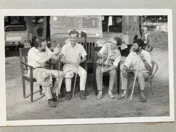 An old black and white photo of four young men sat in chairs with walking sticks in front of Oxfam branded jeeps in Bihar, India.