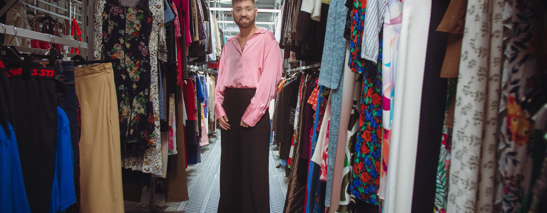 Conor 'The boy in the dress' at the oxfam warehouse at Milton point, wearing a satin pink shirt, and standing in the middle of two rows of clothing