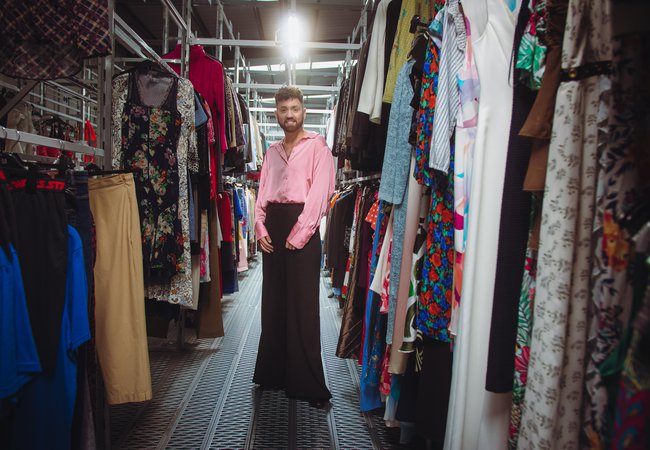 Conor 'The boy in the dress' at the oxfam warehouse at Milton point, wearing a satin pink shirt, and standing in the middle of two rows of clothing