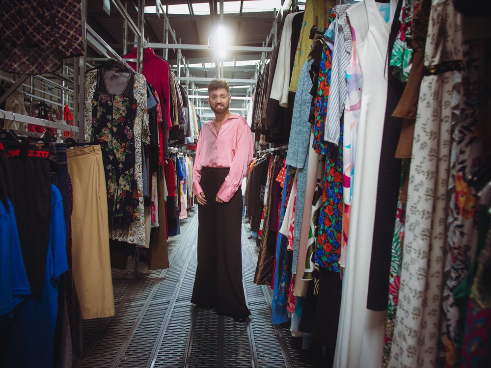 Conor 'The boy in the dress' at the oxfam warehouse at Milton point, wearing a satin pink shirt, and standing in the middle of two rows of clothing