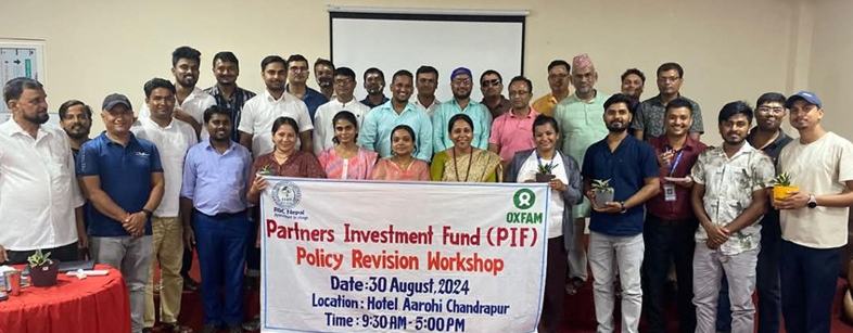 A group of men and women in a hotel function room in Chandrapur, Nepal standing around a white banner promoting the Partners Investment Fund and the Policy Revision Workshop on 30 August 2024.