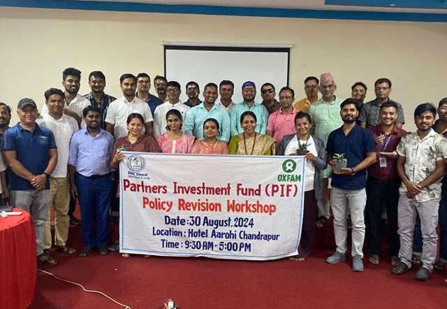 A group of men and women in a hotel function room in Chandrapur, Nepal standing around a white banner promoting the Partners Investment Fund and the Policy Revision Workshop on 30 August 2024.
