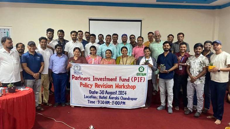 A group of men and women in a hotel function room in Chandrapur, Nepal standing around a white banner promoting the Partners Investment Fund and the Policy Revision Workshop on 30 August 2024.