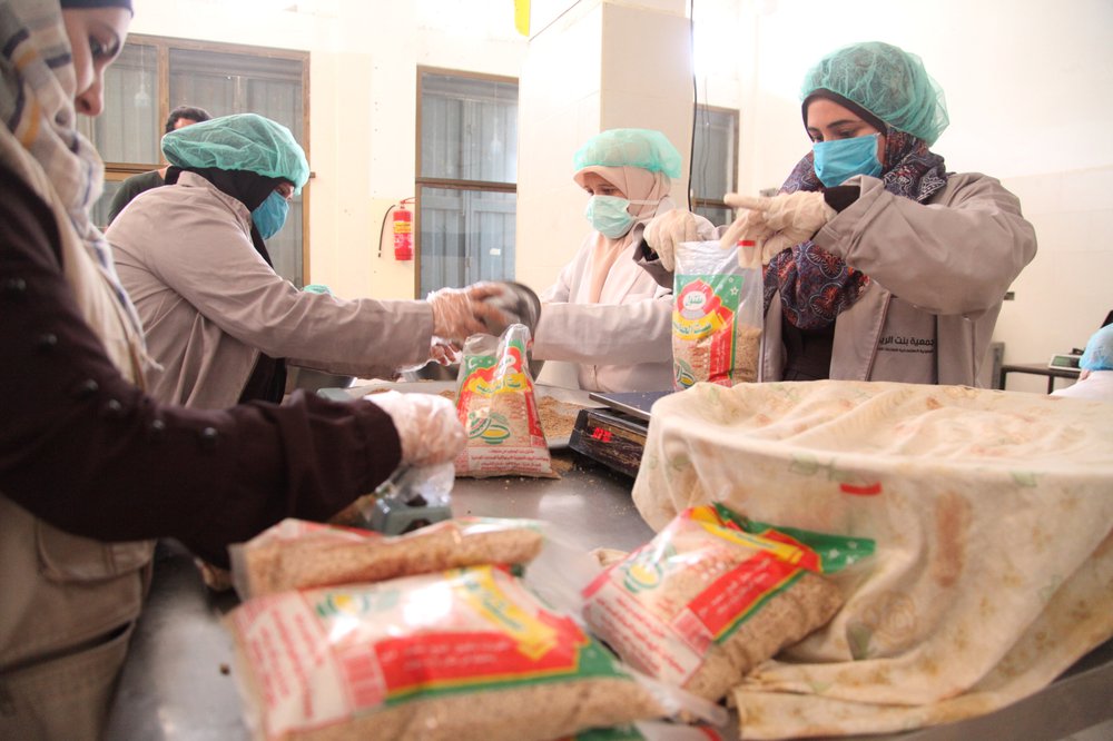 A group of women wearing blue hairnets and facemasks weigh and package food into plastic bags with green red and yellow on them.