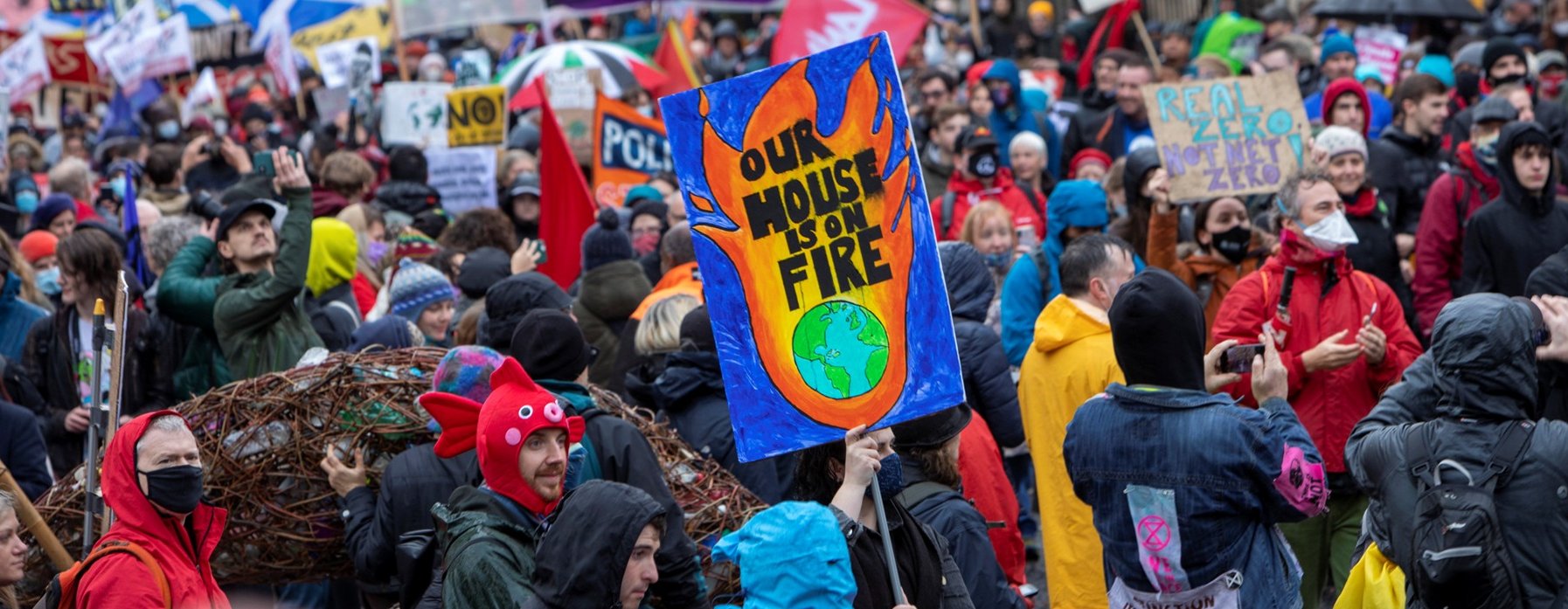 A large crowd of people participate in a climate protest, with one prominent sign showing flames around Earth that reads, "OUR HOUSE IS ON FIRE."