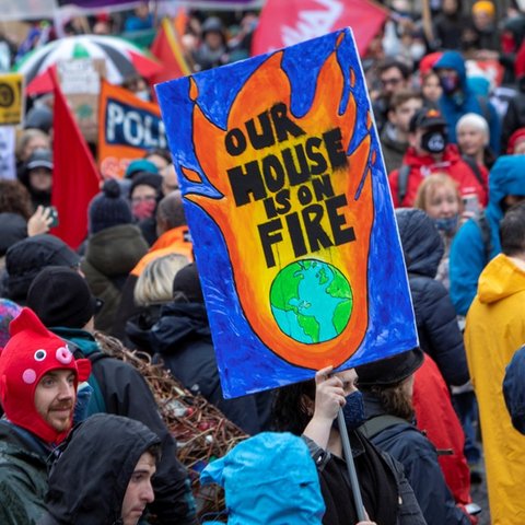 A large crowd of people participate in a climate protest, with one prominent sign showing flames around Earth that reads, "OUR HOUSE IS ON FIRE."