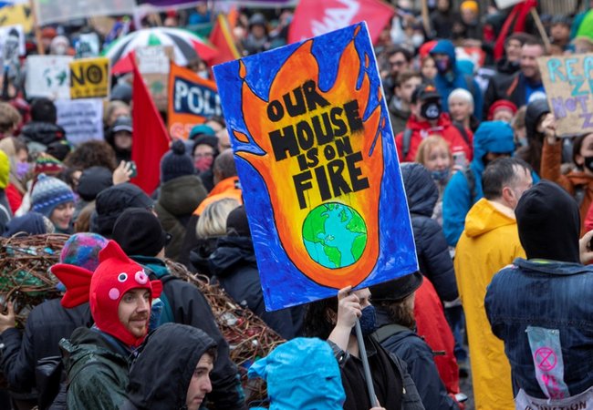 A large crowd of people participate in a climate protest, with one prominent sign showing flames around Earth that reads, "OUR HOUSE IS ON FIRE."