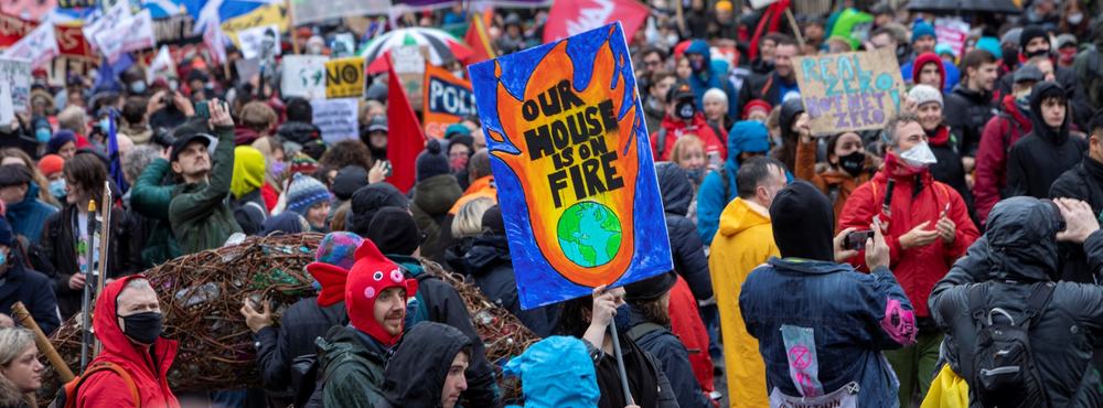 A large crowd of people participate in a climate protest, with one prominent sign showing flames around Earth that reads, "OUR HOUSE IS ON FIRE."