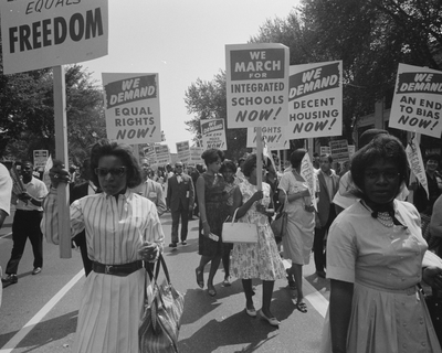 A black-and-white photo of a civil rights march, with people holding signs demanding equal rights, integrated schools, decent housing, and an end to bias. Women and men walk together in the street carrying protest placards.