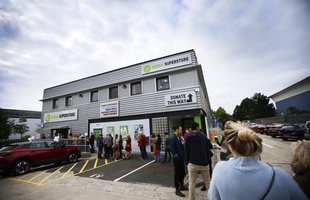 A queue of people stand outside the decorated new Oxfam Superstore, a grey warehouse looking building in Oxford