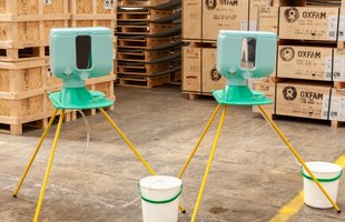 Two fully assembled Handwashing stands in the Oxfam warehouse with boxes stamped with the Oxfam logo that have water tank components in them in the background.