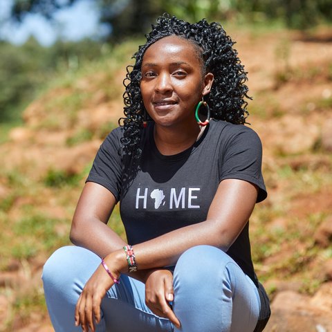 Elizabeth crouches down to pose outside smiling. She's wearing a T-shirt with Africa on it that says 'home' above it.