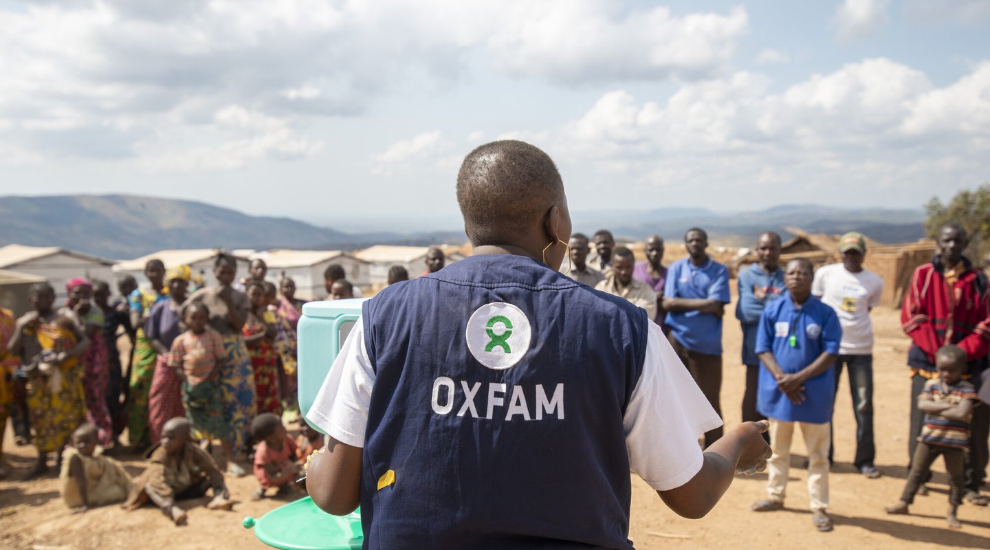 Pascaline has a shaved head and she stands wearing an Oxfam vest with her back to the camera as she talks to community members.