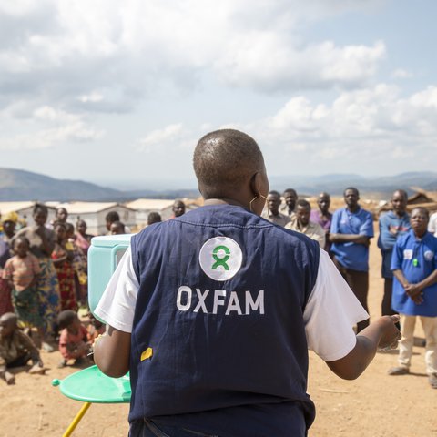 Pascaline has a shaved head and she stands wearing an Oxfam vest with her back to the camera as she talks to community members.