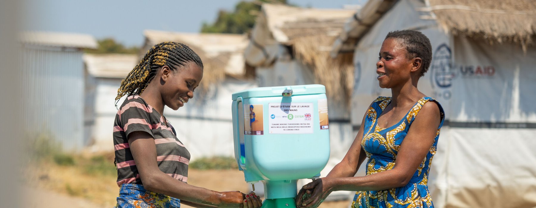 Two young Congolese women wash their hands at a bright blue Oxfam handwashing station on a tripod.