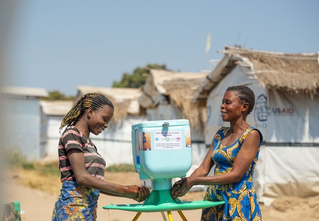 Two young Congolese women wash their hands at a bright blue Oxfam handwashing station on a tripod.