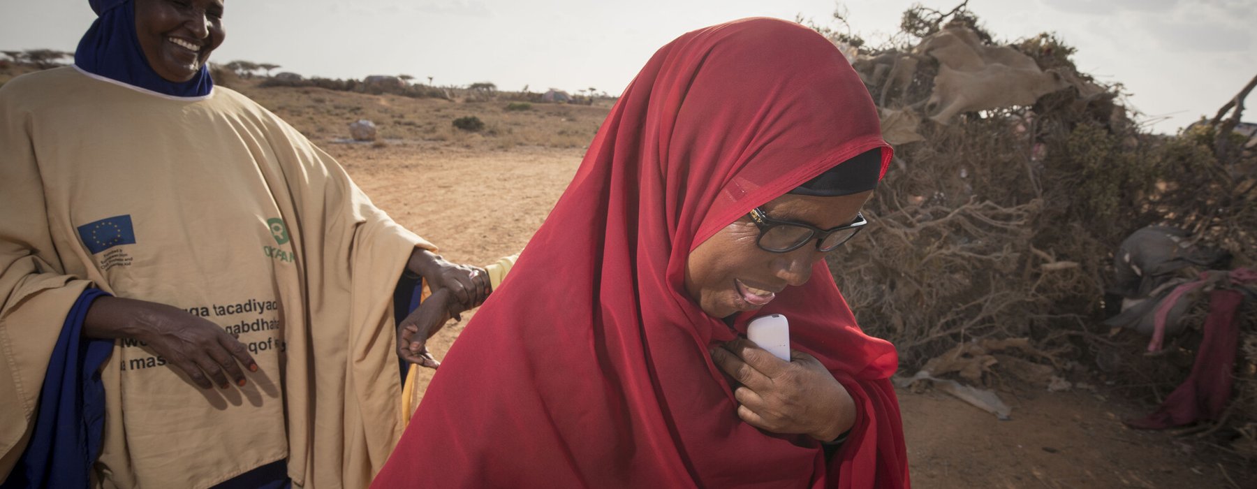Middle aged Somalian Ibaado wears a dark red hijab and smiles as she walks along the ground with a colleague who has a cobalt blue hijab and wears an Oxfam jacket