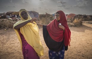 Two women stand in a hot, dusty internally displaced persons camp with tents in the background. They are both over 40 and one wears a yellow hijab and the other a red hijab.