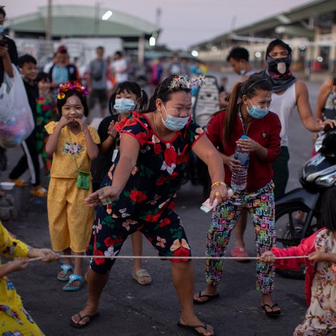 A rope is held out and children wearing masks and with flower garlands in their hair take part in tug of war with adults keeping score and smiling with their eyes.