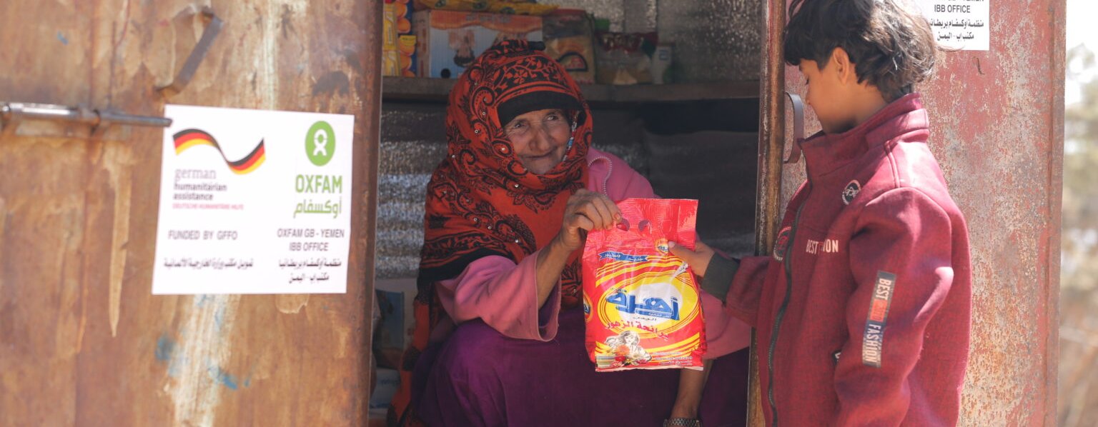 Razigah is sat in the doorway of her small grocery hut handing a red packet to a young boy.